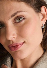 Silverörhängen med böljande design, prydda med små klara stenar. Närbild på ett ansikte, som visar slät hud och mjuk läppfärg.