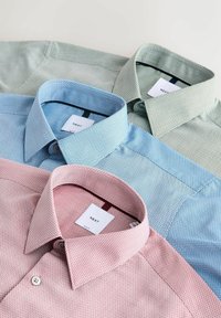 Trois chemises habillées pour hommes soigneusement pliées en rose pastel, bleu et vert avec de fins motifs géométriques et des étiquettes de marque visibles à l'intérieur des cols.