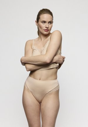 Donna con biancheria intima color beige con rifiniture in pizzo e camicia di raso, con le braccia incrociate, in piedi davanti a uno sfondo grigio chiaro uniforme.