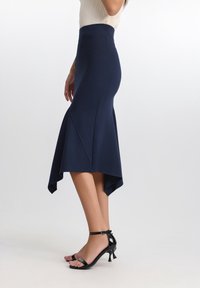 Marinblå midikjol i stretchigt material, med hög midja, utsvängd design och asymmetrisk fåll med spetsiga ändar. Svarta remsandaler.