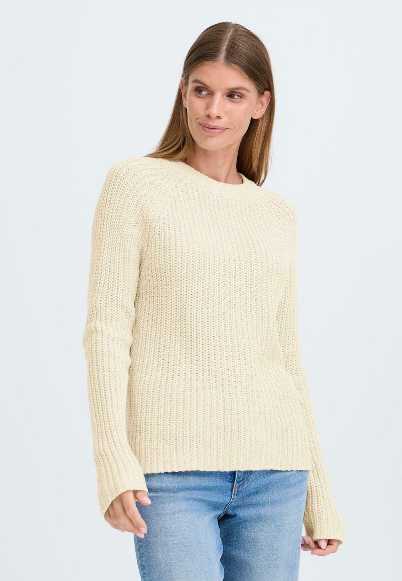 Lys beigefarget strikkegenser med ribbet struktur, rund halsing og lange ermer med forlengede mansjetter, parret med blå jeans.