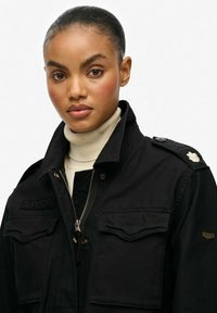 Chaqueta negra con cremallera, que cuenta con dos bolsillos en el pecho, logo bordado "SUPERDRY", hombreras y un pequeño acento floral.
