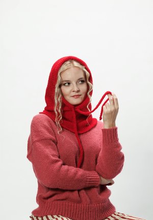 Czerwona dzianinowa czapka z regulowanymi trokami noszona na różowym, oversize'owym swetrze. Materiał jest teksturowany i przytulny, zwiększając ciepło i komfort.