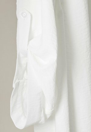 Camicia bianca in tessuto testurizzato con polsini abbottonati. La manica è leggermente arrotolata, mostrando il materiale morbido e leggero.