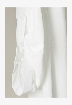 Camicia bianca in tessuto testurizzato con polsini abbottonati. La manica è leggermente arrotolata, mostrando il materiale morbido e leggero.