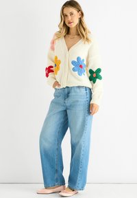 Cream cardigan with colorful floral appliqués, featuring a V-neck and buttons, paired with light blue wide-leg jeans and pink flats.