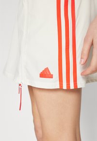 Hvite atletiske shorts med oransje vertikale striper og en oransje Adidas-logo nederst til venstre. Inkluderer en glidelås med en rød pull-tab.