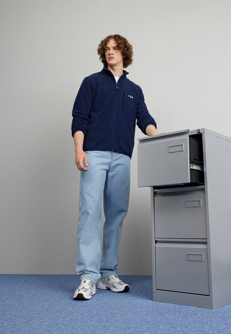 Granatowa polarowa kurtka z suwakiem z detalami kieszeni, jasnoniebieskie dżinsy i białe buty sportowe obok szarej szafy na dokumenty.