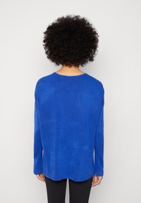 Personne aux cheveux bouclés et foncés, portant un pull à manches longues bleu vif et un pantalon noir, vue de dos sur un fond clair uni.