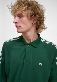 Blond person med krøllete hår som har på seg gullfargede runde øredobber og en mørkegrønn skjorte med krage og hvite sikksakkstriper på skuldrene.