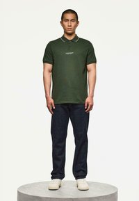 Muž stojí na kulaté platformě, oblečený v tmavě zeleném polo tričku s textem, tmavých džínách a bílých teniskách, na jednobarevném světlém pozadí.