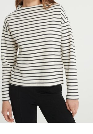 Femme portant une chemise blanche à manches longues avec des rayures horizontales noires et un pantalon noir, debout devant un fond uni.