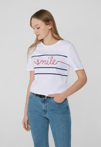 Camiseta de algodón blanca de manga corta, con un texto de "sonrisa" en rojo, flanqueado por dos franjas horizontales en azul marino. Combinada con pantalones vaqueros azules.