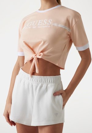 Kvinde der bærer en ferskenfarvet croppet tie-front Guess t-shirt og hvide shorts med elastik i taljen og logo, hånd i lommen, stående mod en hvid baggrund.
