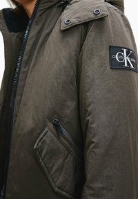 Kurtka puchowa w oliwkowym zielonym kolorze o gładkiej, teksturowanej powierzchni; posiada stójkę, kieszeń na zamek i naszywkę z logo na ramieniu.