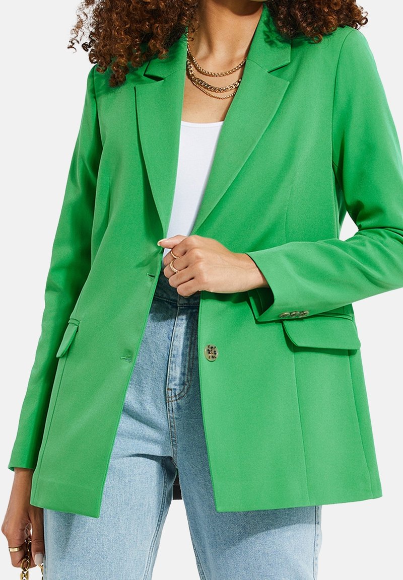 Femme portant un blazer vert vif sur un haut blanc et un jean bleu, accessoirisée avec des colliers et des bagues en or, sur fond blanc.