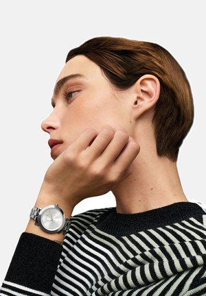 Perfil lateral de una persona con el cabello corto y castaño que lleva un suéter a rayas blancas y negras y un reloj de pulsera plateado en su muñeca izquierda.