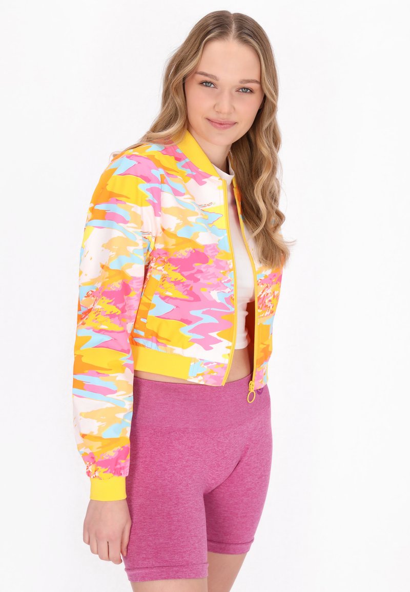 Kleurvolle bomberjack met gewaagde roze, gele en blauwe patronen, met elastische manchetten en tailleband. Gepaarde met roze shorts.