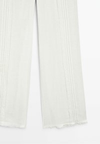 Lekka, kremowa para spodni o luźnym kroju. Posiada wyszywane pionowe detale oraz postrzępione krawędzie dołu dla dodania tekstury.