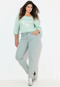 Top de manga larga verde claro con texto plateado, combinado con jeans ajustados gris claro y botas de tobillo plateadas. Textura suave y diseño casual.