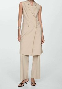 Blazer sans manches beige à double boutonnage, associé à un pantalon à jambes larges. Tissu lisse avec une coupe ajustée. Sandales à talons noires avec des accents.