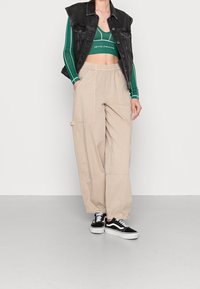 Beige wijde cargobroek met grote zakken, gecombineerd met een donker denimvest en een groene crop top met strepen. Zwart-witte sneakers maken de look compleet.
