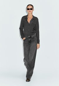 Dunkelgraues Hemd mit Knopfverschluss und zwei Brusttaschen, kombiniert mit hochtaillierten schwarzen Jeans. Das Model trägt schwarze Sonnenbrillen und braune Stiefel.