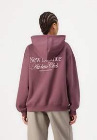 Sudadera con capucha en color malva, que presenta una capucha y un gran texto gráfico blanco en la parte trasera. Puños y dobladillo de canalé con un diseño de corte relajado.
