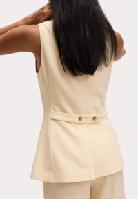 Chaleco sin mangas beige con un corte entallado, detalles de botones en la parte trasera y una textura suave y estructurada.