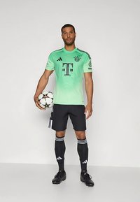 Ποδόσφαιρική φανέλα με πράσινο βαθμιδωτό χρώμα και μαύρο σορτς, με το λογότυπο και τρεις μαύρες ρίγες στα μανίκια και τις κάλτσες, κρατώντας μια μπάλα ποδοσφαίρου.