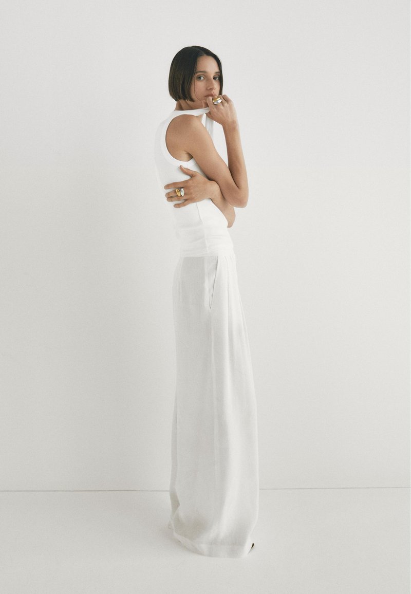 Biały dopasowany top na ramiączkach w połączeniu z luźną, wysoką spódnicą z białego lnu. Dodatki to złote pierścionki na palcach. Neutralne tło.