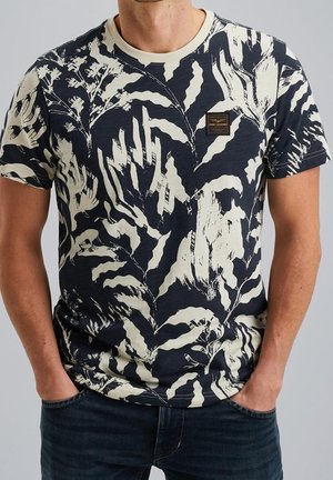 Homme portant un t-shirt bleu marine avec des motifs abstraits de feuilles blanches et un petit patch logo rectangulaire sur la poitrine, les mains dans les poches.
