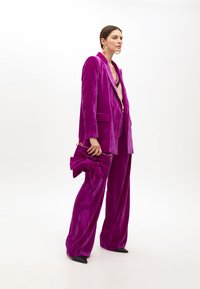 Welurowy zestaw składający się z długiego żakietu i spodni o szerokich nogawkach w intensywnym purpurowym kolorze. Zawiera kopertówkę z kokardą oraz czarne, spiczaste buty.