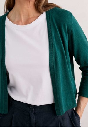 Femme portant un T-shirt blanc à col rond sous un cardigan court vert foncé et un pantalon foncé taille haute, les mains dans les poches.