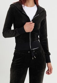 Sweat à capuche zippé en velours noir avec manches longues et poignets côtelés, assorti d'un pantalon avec cordon de serrage. Texture lisse, coupe décontractée.