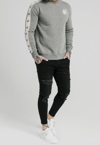 Mężczyzna w szarym dresie z logo na piersi, w czarnych, przetartych skinny jeansach i białych trampkach.