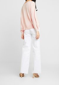 Jasnoróżowy sweter z balonowymi rękawami w połączeniu z wysokim stanem białych, szerokich spodni dżinsowych. Modelka nosi beżowe, wysokie obcasy.