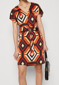 Šaty s krátkým rukávem a véčkovým výstřihem, s vícerozměrným geometrickým vzorem v červené, oranžové, žluté, černé a bílé barvě. Detaily vázaného pasu.