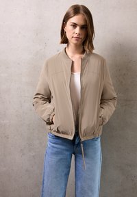 Veste bomber beige avec fermeture éclair à l'avant, col côtelé, poignets élastiques et ourlet. Associée à un jean en denim droit bleu clair.