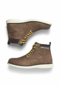 Bottines en cuir marron avec des lacets jaunes, semelles en caoutchouc blanches et étiquette logo noir et blanc sur le col. Fini texturé, design robuste.