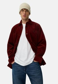 Ung mand iført beige strikket beanie, burgunder fløjlskurt over hvid t-shirt og blå jeans med hænderne i lommerne mod en ensfarvet baggrund.