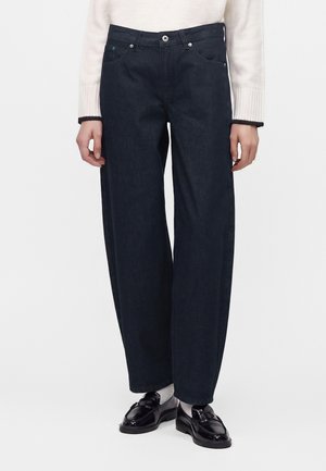 Person iført mørke vide jeans, hvide sokker, sorte skinnende loafers og en hvid langærmet top, stående mod en ensfarvet baggrund.