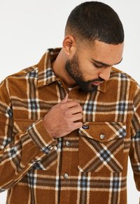 Brown plaid shirt made of soft fabric, featuring blue and white accents, a button-up front, and a single chest pocket with a flap.