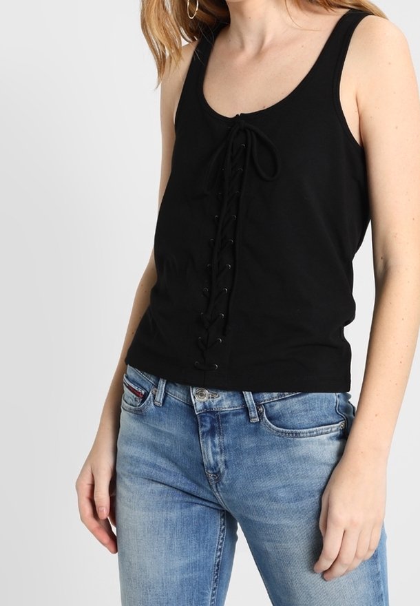 Débardeur noir avec un détail lacé à l'avant, fabriqué en tissu doux. Porté avec un jean denim bleu clair. Design simple, coupe sans manches.