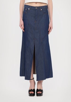 Femme portant une jupe longue en denim bleu foncé taille haute avec une fente à l'avant, associée à des sandales à plateforme noires avec des brides à la cheville.