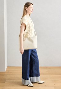 Dziergany biały top z wzorem warkocza, zestawiony z szerokimi dżinsami o szerokich nogawkach z kontrastowymi mankietami oraz teksturowaną, jasnobeżową torebką.