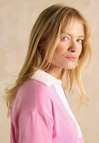 Top tricotat roz cu guler alb, având o croială relaxată și o textură moale. Părul cade pe umeri pe un fundal neutru.