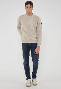 Giovane uomo in piedi su uno sfondo bianco, vestito con un maglione beige, jeans blu scuro e sneakers bianche con dettagli blu e beige.
