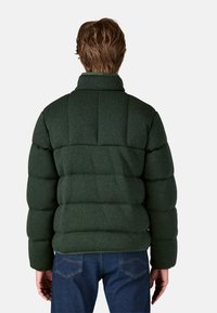 Giacca a vento verde con colletto alto, realizzata in un tessuto texturizzato, con cuciture verticali evidenti e un design imbottito. Indossata con jeans blu.