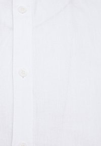 Camicia bianca con bottoni a pressione, realizzata in un tessuto testurizzato, con bottoni bianchi allineati centralmente davanti e cuciture sottili lungo i bordi.